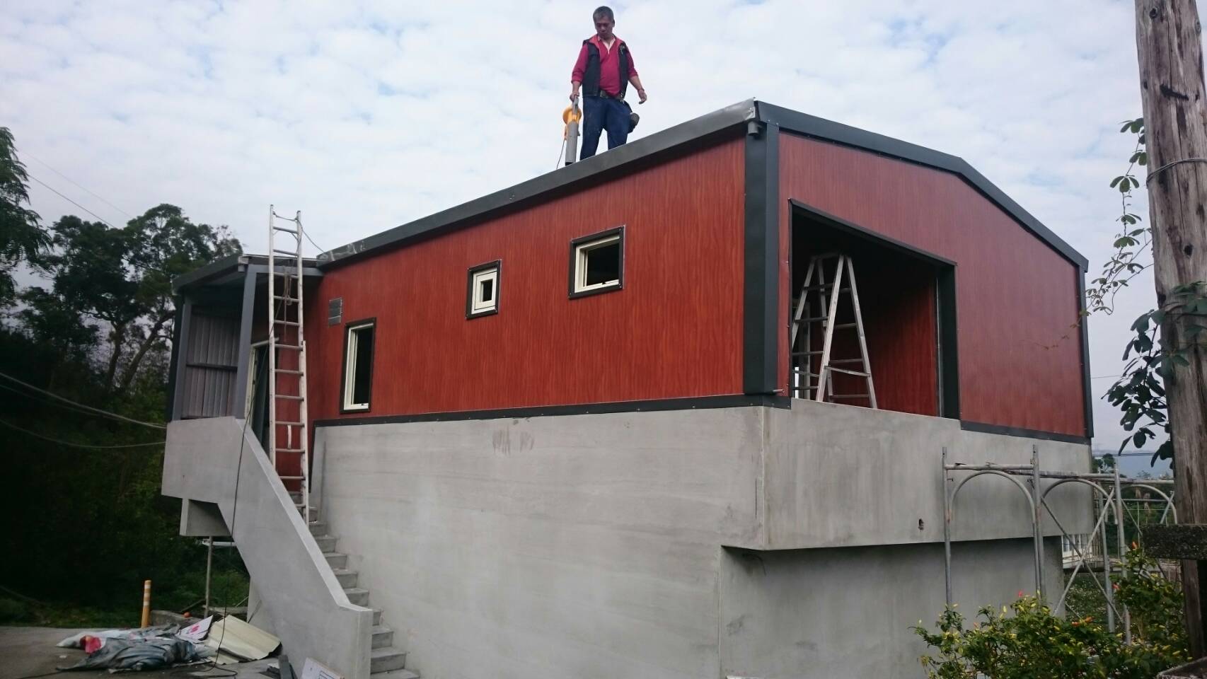 1) 照片顯示一座兩層樓的建築物，建築物外牆為紅色木板，上面有一個人站在屋頂上，建築物外觀看起來很新，旁邊有綠色的植物